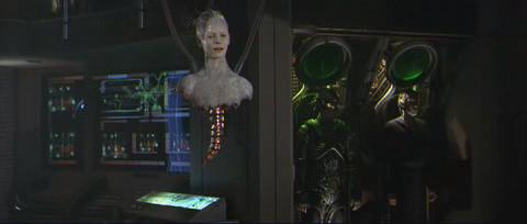 Video clip from the film, showing the disembodied head and shoulders of a woman being  lowered into a tight black suit by cables. The woman has pale, mottled skin and wires protruding from the back of her head; a mechanical spine hangs from the rest of her upper body.
